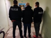 Na zdjęciu widoczni policjanci ubrani po cywilnemu z założonymi na plecach kamizelkami taktycznymi z napisem Policja. Policjanci prowadzą &quot;pod ręce&quot; zatrzymanego mężczyznę ubranego w ciemną kurtkę. Zdjęcie wykonane w jednostce policji.