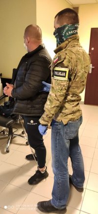 Policjant po cywilnemu ubrany w kurtkę w moro trzyma podejrzanego. Zdjęcie wykonane w pomieszczeniu policji. Policjant i zatrzymany mają na twarzach maseczki. Zatrzymany ma założone na ręce i nogi kajdanki (prowadnice)