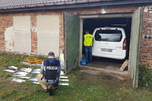 dziupla samochodowa zlikwidowana przez policjantów, widoczny tył białego pojazdu i stojący obok funkcjonariusz w żółtej kamizelce, przed budynkiem na trawie leżą rozłożone tablice rejestracyjne przy których kuca funkcjonariusz w czarnej kamizelce z napisem policja