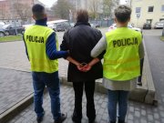 Na zdjęciu widocznie dwaj policjanci po cywilnemu, którzy mają założone kamizelki odblaskowe żółtego koloru z czarnym napisem policja. Policjanci trzymają pod ręce mężczyznę ubranego w czrny płaszcz i ciemne spodnie, który ma założone kajdanki na ręce trzymane z tyłu. W dalszym planie widoczny jest parking samochodowy.