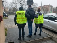 Zdjęcie jest zrobione na dziedzińcu Komendy Miejskiej Policji w Łodzi, widzimy na nim zatrzymanego ubranego w ciemną kurtkę, dźinsy. Jest wysoki, szczupłej budowy ciała, ma ciemne włosy. Ręce ma skute kajdankami. Po jego lewej stronie trzyma go policjant ubrany w żółtą kamizelkę policyjna, a po prawej stronie stoi policjantka, ubrana również w żółtą kamizelkę policyjną