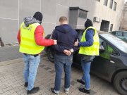 Na zdjęciu widzimy zatrzymanego, który jest prowadzony do samochodu przez policjanta i policjantkę. Zdjęcie jest zrobione na dziedzińcu Komendy Miejskiej Policji, sprawca ubrany jest w szarą bluzę z kapturem oraz dżinsy, natomiast policjanci maja ubrane zielone kamizelki policyjne z napisem Policja.