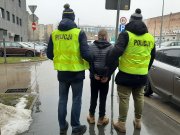 Dwóch policjantów ubranych w czapki oraz w zielone policyjne kamizelki  z napisem Policja prowadzi zatrzymanego, ubranego w czarną kurtkę. Mężczyzna jest niskiego wzrostu. Wszyscy są odwróceni tyłem, mężczyzna ma założone z tyłu kajdanki.