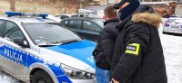 Policjant, ubrany w czarna kurtkę zimową, kominiarkę, opaskę z napisem policja oraz czarną czapkę bejsbolówkę trzyma zatrzymanego. Mężczyzna ubrany jest w czarną kurtkę i dżinsy, ma ciemne włosy. Obaj stoją przy policyjnym radiowozie.