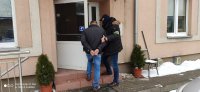 Policjant wraz z zatrzymanym wchodzą do II komisariatu, mężczyzna ma założone kajdanki na rękach, policjant otwiera mu białe, oszklone drzwi. Przy wejściu stoją w donicach dwie tuje, budynek komisariatu jest koloru brudno-różowego.