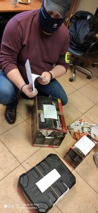 Policjant po cywilnemu, ubrany w kominiarkę oraz czarną czapkę bejsbolową kuca na podłodze i ogląda odzyskane elektronarzędzia.
