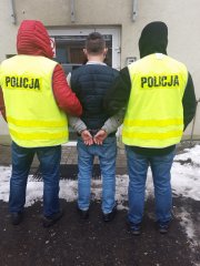 Dwóch policjantów, ubranych w kurtki zimowe- czerwoną i czarną, ma założone kaptury na głowie oraz żółte kamizelki z napisem Policja. Policjanci wraz z zatrzymanym są odwróceni tyłem do aparatu. Zatrzymany mężczyzna  ma z tyłu założone kajdanki . Ubrany jest w szarą bluzę, w granatową kamizelkę oraz niebieskie spodnie. Zdjęcie jest zrobione przed komisariatem.