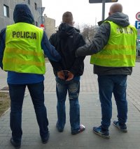 Mężczyzna stoi przy policyjnym areszcie , po jego obu stronach stoją policjanci ubrani w policyjne kamizelki. Mężczyzna ma założone kajdanki na ręce trzymane z tyłu, Wszyscy mają na sobie ciemne kurtki zimowe, w tle widać I Komisariat Policji.