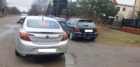 Nieoznakowany radiowóz policyjny, w kolorze srebrnym, na tylnej szybie wyświetla się napis Policja, obok stoją zaparkowane samochody