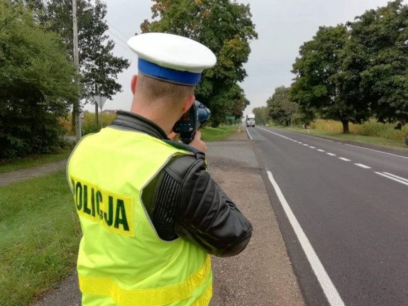 Policjant, ubrany w żółtą kamizelkę policyjną i białą czapkę jest odwrócony tyłem do aparatu i mierzy prędkość pojazdów. Stoi przy drodze asfaltowej, po obu stronach są drzewa.