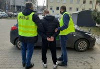 Na zdjęciu widzimy zatrzymanego , który jest prowadzony przez policjantów do cywilnego radiowozu. Policjanci mają zielone kamizelki policyjne . Zatrzymany ma na sobie ciemną, zimową kurtkę z kapturem. Zdjęcie jest zrobione na dziedzińcu Komendy Miejskiej Policji w Łodzi.