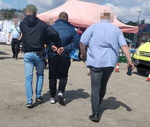 Policjanci prowadzą podejrzanego mężczyznę przez rynek. Mężczyzna ma założone kajdanki na ręce trzymane z tyłu.