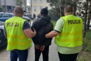 mężczyzna ubrany w czarną kurtkę z kapturem i czarne spodnie jest trzymany po obu stronach przez dwóch policjantów, ubranych w żółte kamizelki policyjne. Zdjęcie jest zrobione na dziedzińcu Komendy Miejskiej Policji.