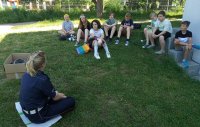 Ośmioro dzieci siedzi na trawie i słuchają co ma do powiedzenia policjantka.