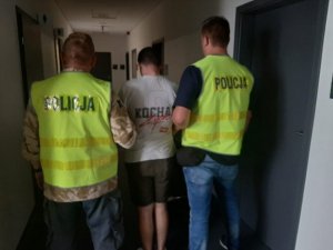 zatrzymany jest na I Komisariacie Policji i jest prowadzony korytarzem przez dwóch policjantów. Ubrany jest w białą koszulkę z napisami i zielone krótkie spodenki.