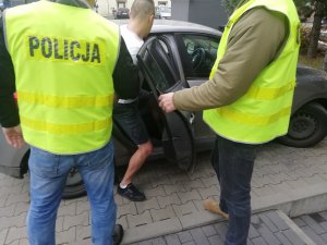 Nieumundurowani policjanci z zatrzymanym mężczyzną. Mężczyzna wsiada do nieoznakowanego radiowozu Policji.