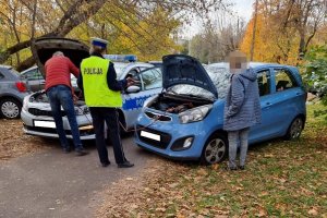 Miejsce unieruchomienia samochodu, policjantka wraz mężczyzną podpinają kable. Obok stoi właścicielka auta.