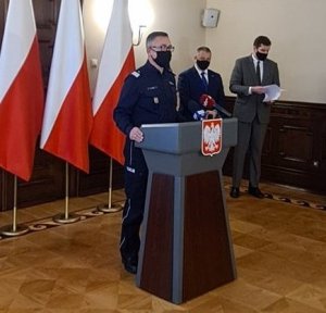 generał policji stoi za mównicą i zabiera głos, w tle dwóch mężczyzn tj. wojewoda i kurator.obok trzy flagi biało-czerwone