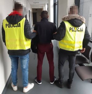 Korytarz komisariatu. Dwaj policjanci w żółtych kamizelkach odblaskowych prowadzą zatrzymanego mężczyznę.