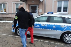 Policjant na tle radiowozu prowadzi zatrzymanego w kajdankach sprawcę kradzieży plecaka.