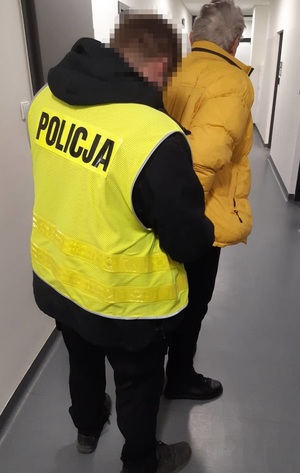 Policjant wraz z zatrzymanym w budynku Komendy. Mężczyzna ma założoną żółta kurtkę i kajdanki na ręce trzymane z tyłu.