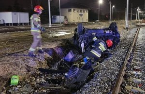 Rozbity pojazd leżący na torach przy nim pracujący strażacy.