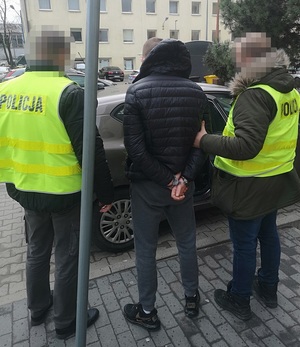 Zatrzymany z założonymi kajdankami na ręce trzymane z tyłu w asyście policjantów.