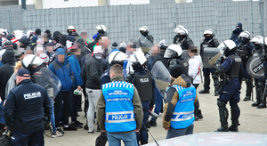 Zabezpieczenie meczu, policjanci eskortują kibiców gości na teren stadionu. Wśród funkcjonariuszy sa nieumundurowani w kamizelkach z napisem policja, zespół antykonfliktowy.