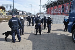 Policjanci nadzorują wejście na stadion kibiców przyjezdnych, w tym patrol z psem służbowym. Obok stoją zaparkowane specjalistyczne radiowozy.