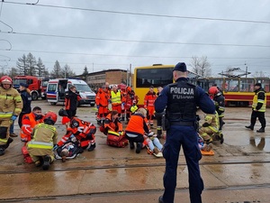 Policjant zabezpiecza miejsce zdarzenia, ratownicy udzielają pomocy ofiarom katastrofy.