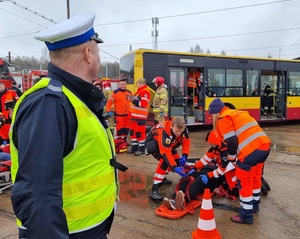 Policjant zabezpiecza miejsce zdarzenia, ratownicy udzielają pomocy ofiarom katastrofy.