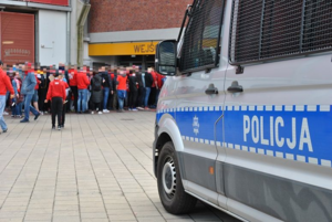 Radiowóz policyjny przed wejściem na stadion przed którym stoją kibice.