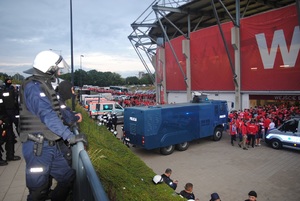 Kibice gospodarzy wchodzą na stadion. Przed wejściem zaparkowany pojazd specjalistyczny z armatka wodną. Wokół policjanci pilnują porządku.
