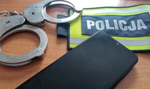 Kajdanki, telefon komórkowy , opaska z napisem POLICJA leżą na blacie biurka.