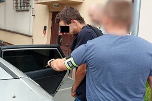 Nieumundurowany policjant wyprowadza podejrzanego z radiowozu. Policjant ubrany jest po cywilnemu, na ramieniu ma opaskę z napisem Policja.