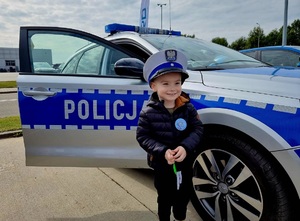 Dziecko ubrane w czapkę policyjną stoi przed oznakowanym radiowozem.