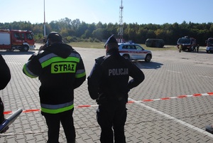 Policjant o praz pracownik Państwowej Straży Pożarnej obserwują teren akcji ratunkowej na lotnisku.