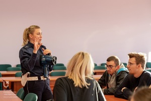 Policjantka rozmawia ze słuchaczami, w dłoni trzyma urządzenie do pomiaru prędkości.