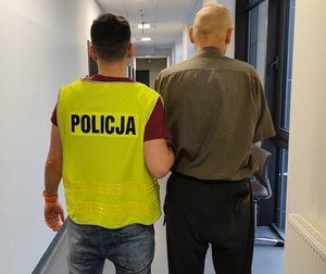 Funkcjonariusz w kamizelce z napisem policja prowadzi korytarzem komisariatu zatrzymanego sprawcę.