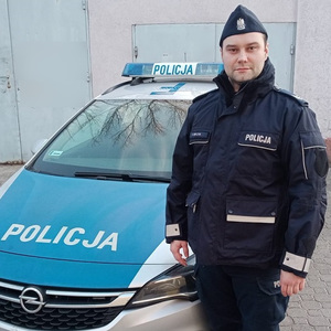 Umundurowany policjant stoi przy oznakowanym radiowozie.