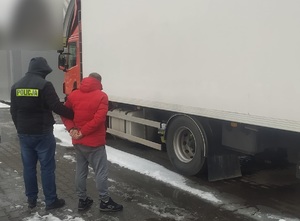 Policjant prowadzi zatrzymanego , przed nimi stoi zaparkowany samochód ciężarowy.