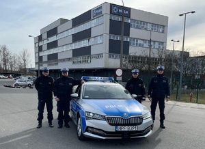 Czterech policjantów stoi po bokach oznakowanego radiowozu.
