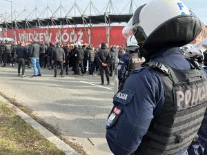 Umundurowani policjanci w kaskach i kamizelkach ochronnych obserwują kibiców stojących przed wejściem na stadion.