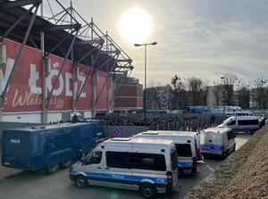 Teren wokół stadionu, na pierwszym planie kilka radiowozów, w tle zgromadzeni przed wejściem kibice oraz pilnujący porządku policjanci.