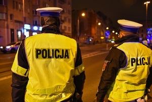 Policjanci stoją przy jezdni obserwują ruch uliczny, zmrok rozświetlają latarnie uliczne.