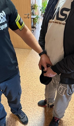 Policjant trzyma zatrzymanego, który ma kajdanki na rękach i nogach.