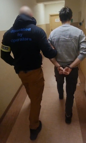 Policjant prowadzi korytarzem zatrzymanego sprawcę, który ma na dłoniach kajdanki.
