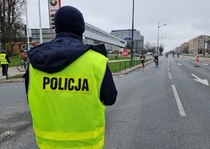 Policjant stoi na jezdni po której wytyczona jest trasa maratonu, w tle biegną zawodnicy.