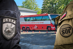 Pomiędzy umundurowanymi funkcjonariuszami służb w tle stoi autobus wykorzystywany do mobilnego pobierania krwi.