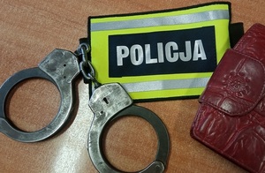 Na stole leżą kajdanki, opaska z napisem policja oraz czerwony portfel.